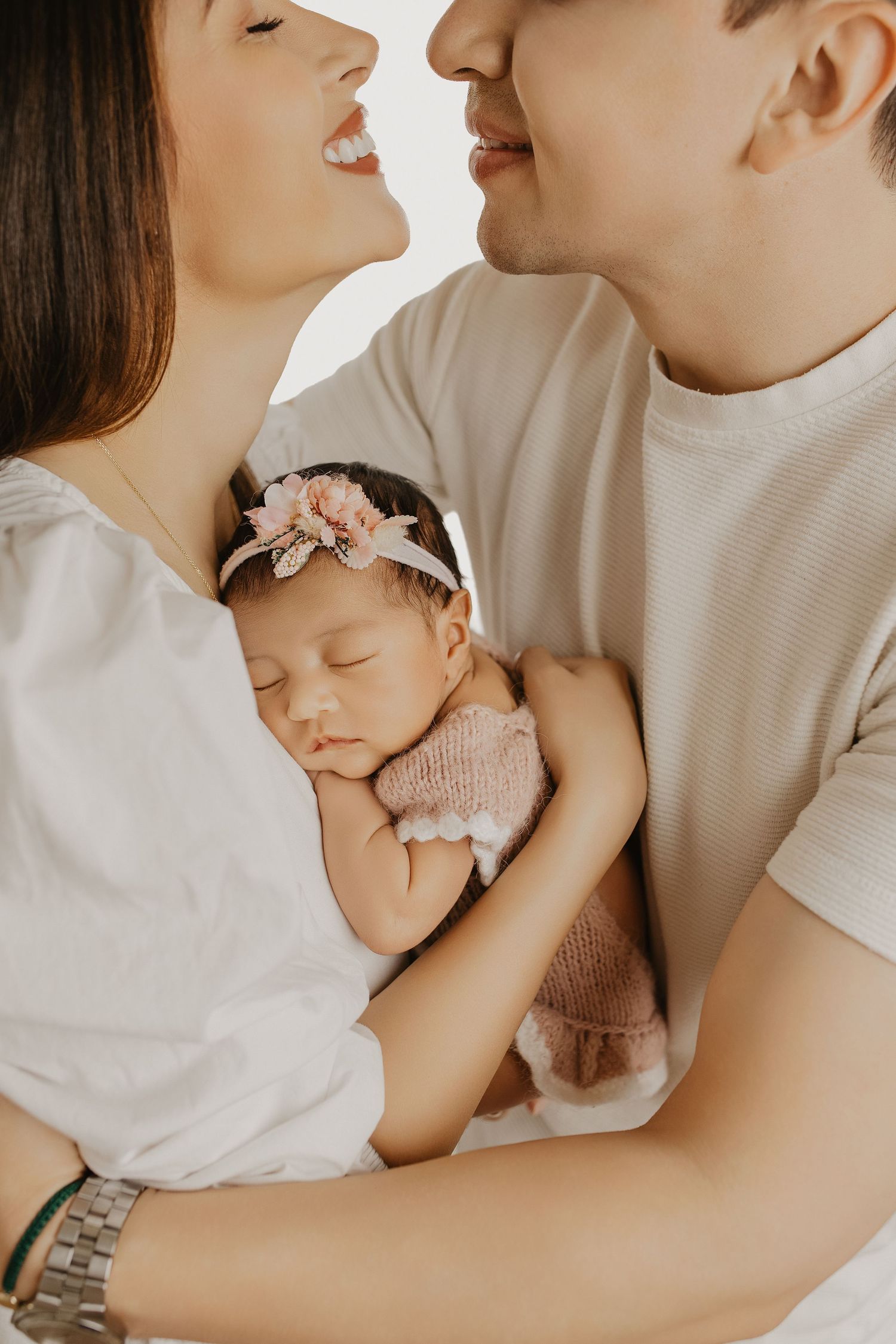 Parents smiling at each other while gently holding their sleeping newborn baby girl wearing a pink floral headband.