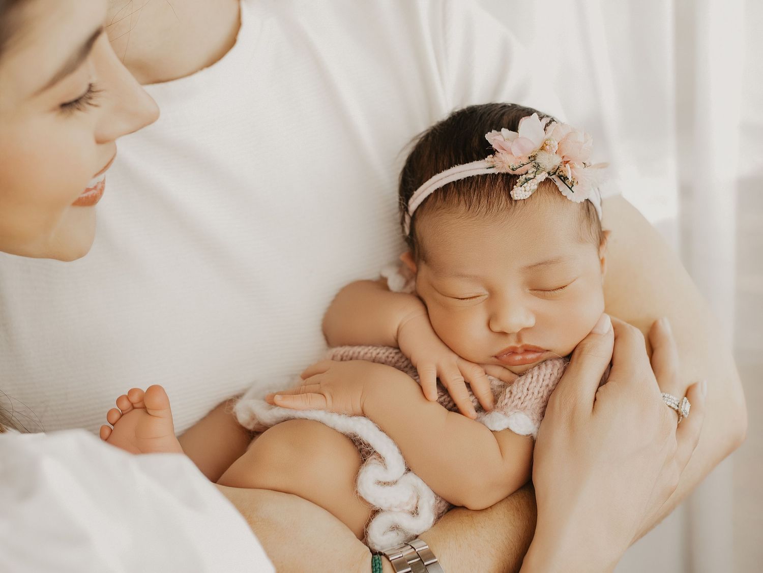 Newborn baby girl with floral headband sleeping peacefully in mother's arms during professional newborn photography session.