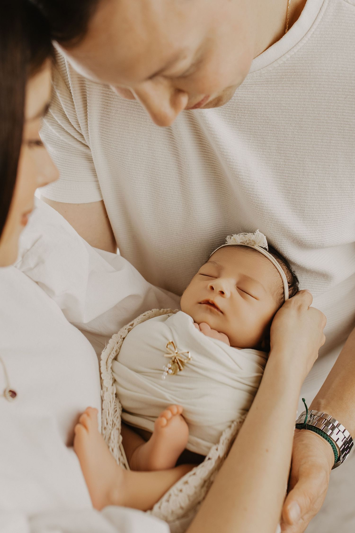 Parents lovingly gaze at their sleeping newborn baby wrapped in white swaddle with gold bow headband in a basket.