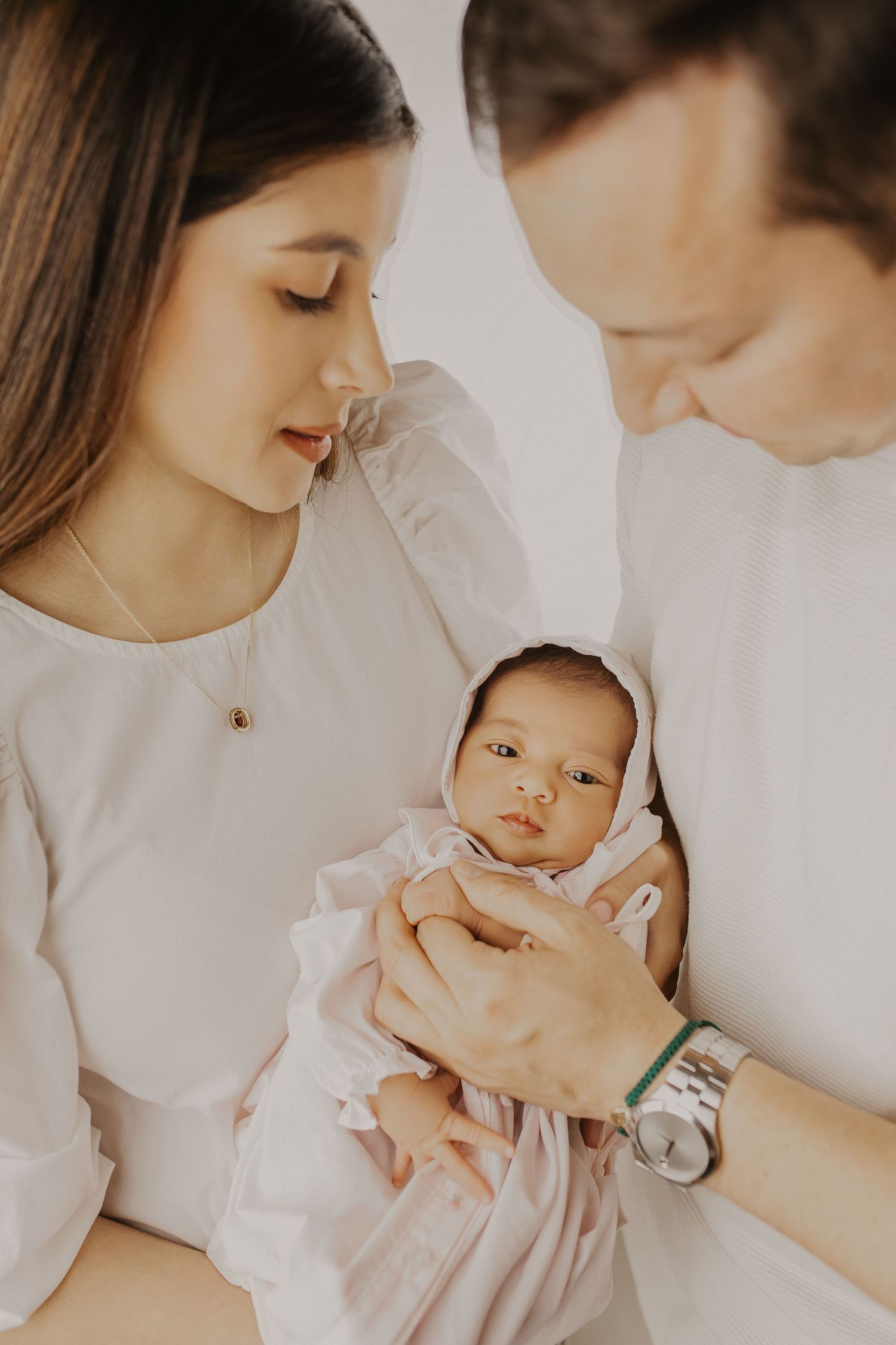 A couple in white clothing tenderly holds their newborn baby wrapped in a soft pink blanket.