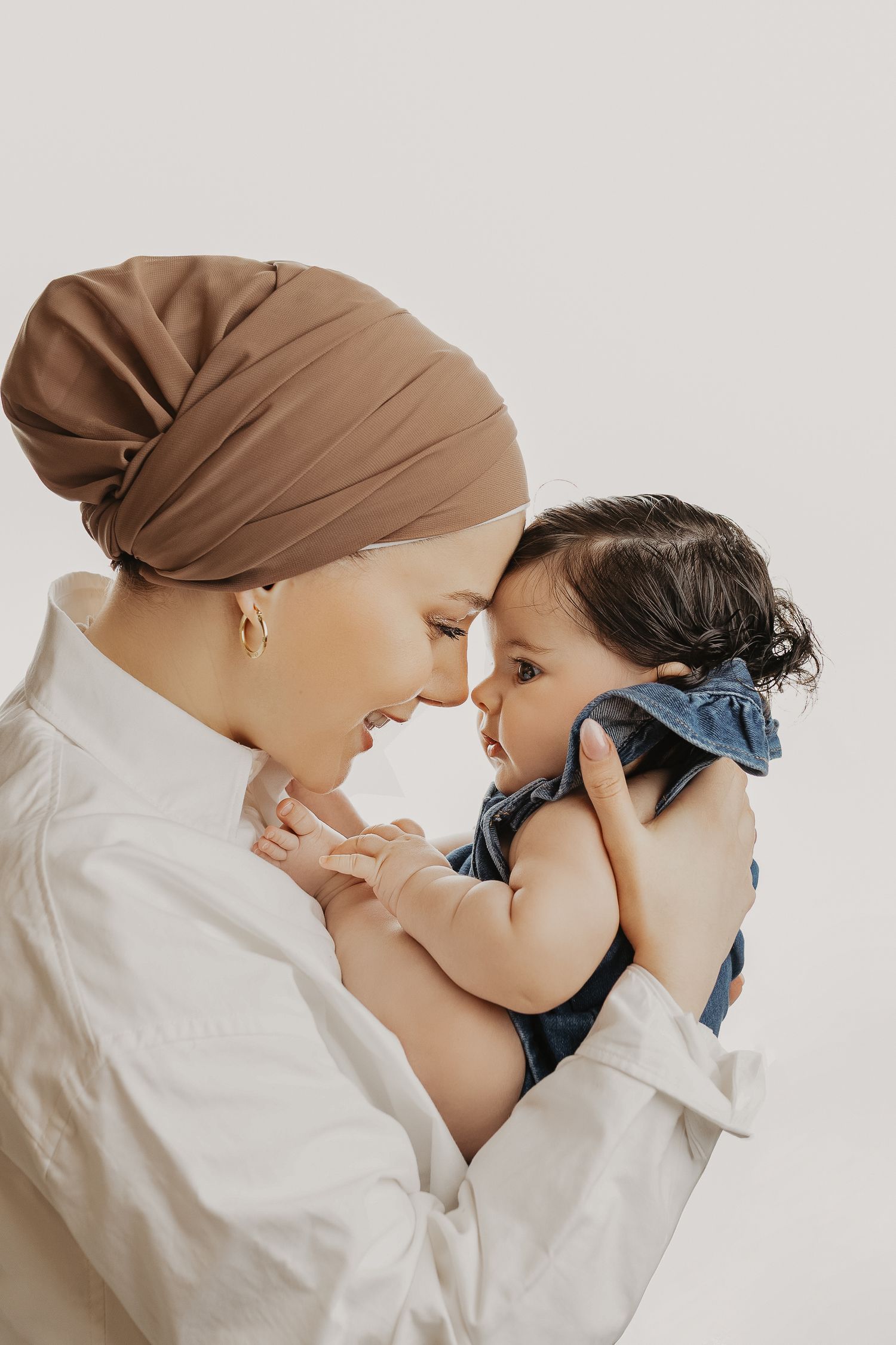 A mother wearing a brown hijab tenderly holds her baby girl in a denim outfit, touching foreheads in a loving moment.