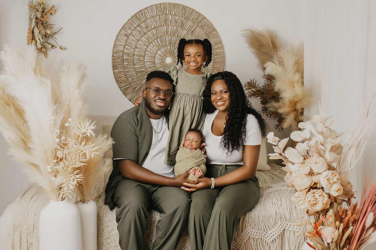 Happy family of four posing in a boho studio, parents holding newborn baby, young daughter standing behind them.