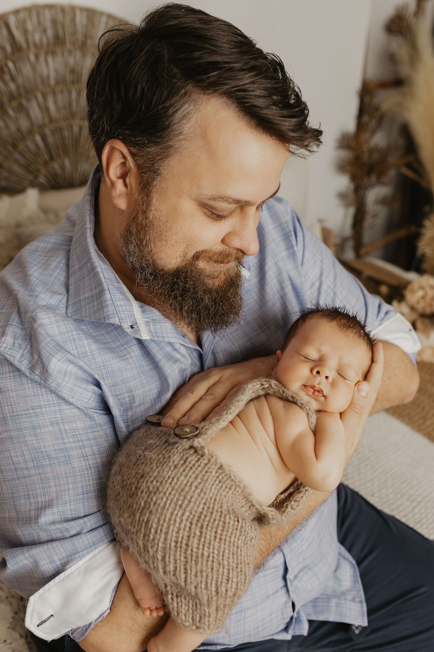 Parent in blue shirt cuddling sleeping baby wearing knit bonnet in warm indoor setting.