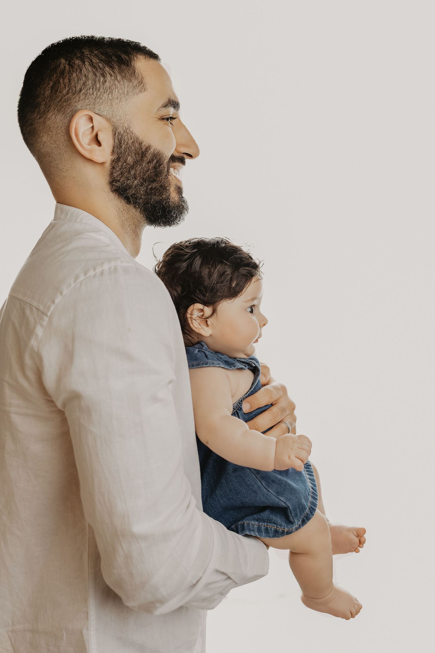 Smiling bearded man in white shirt holding a baby in denim overalls against a light background.