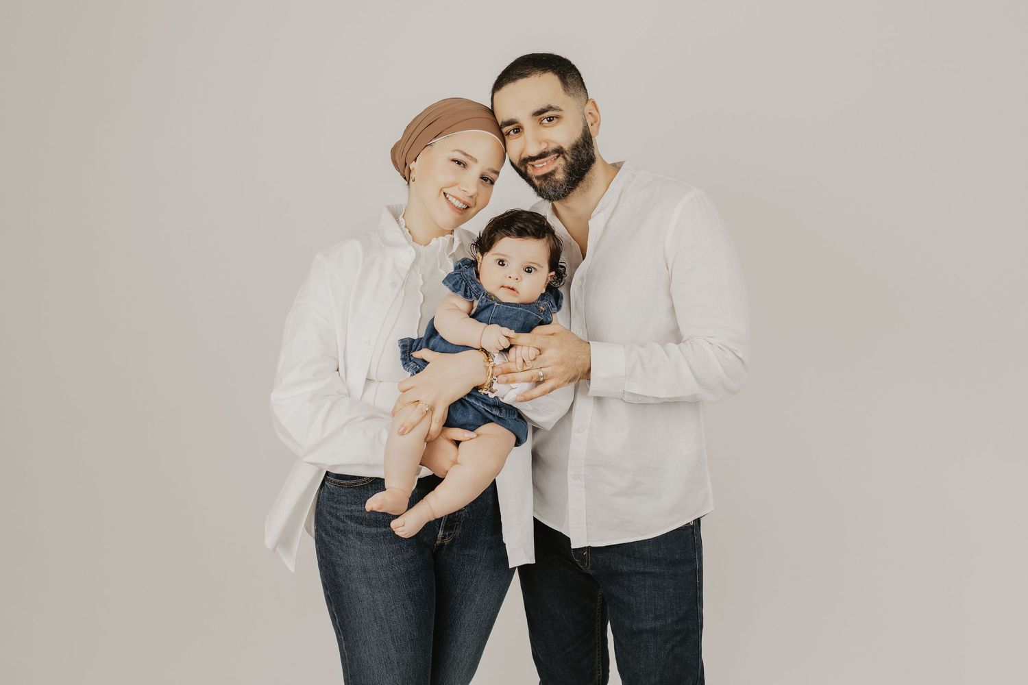 A smiling family of three poses together, with parents in white shirts and jeans holding their baby dressed in denim.