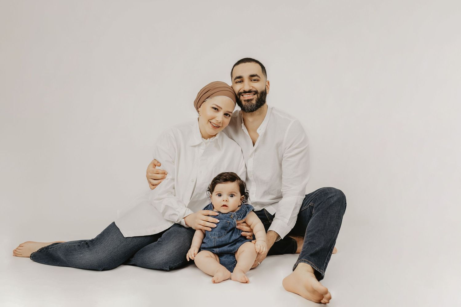 A smiling family of three sits on a white studio floor, parents in white shirts with their baby in a denim outfit.