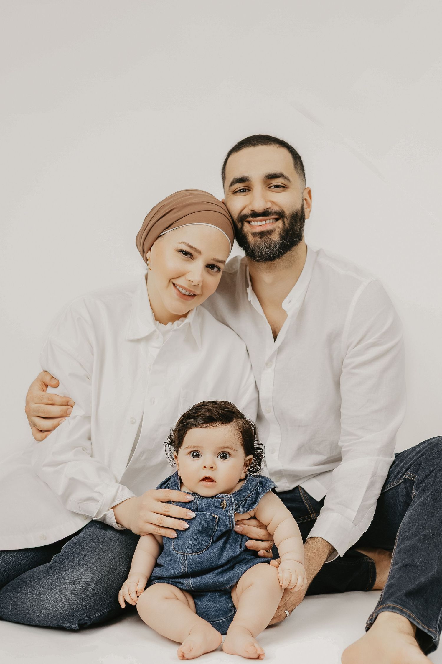 A smiling Muslim family with a woman in hijab, bearded man in white shirts, and their baby girl in denim overalls.
