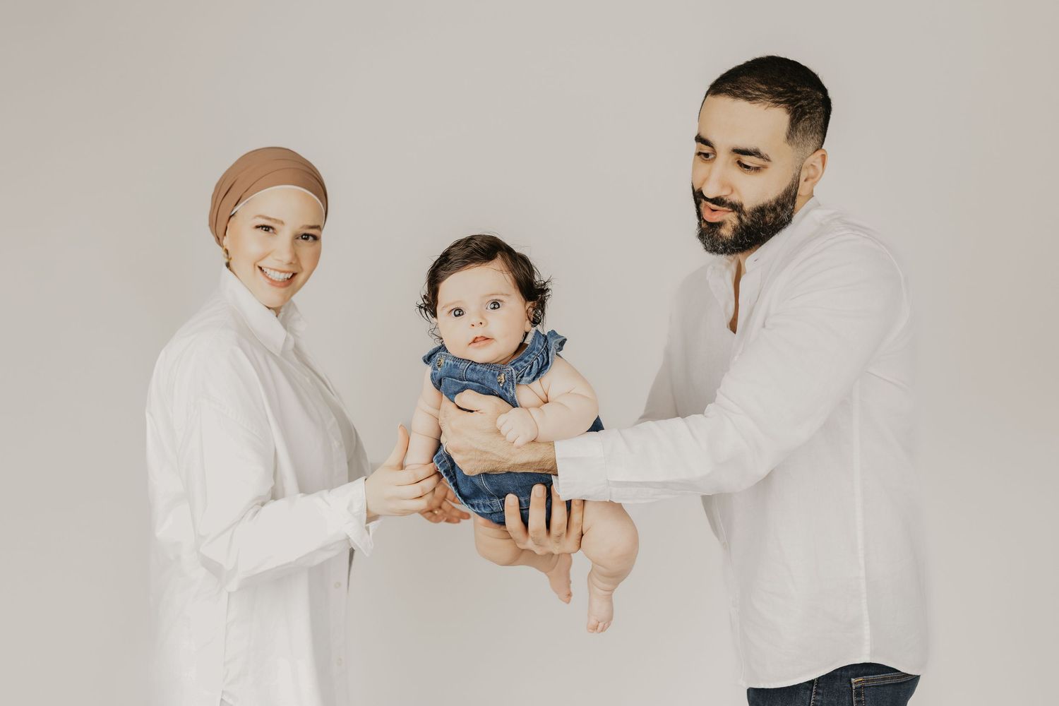 A smiling Muslim family with a mother in hijab and father holding their baby in a bright white studio setting.