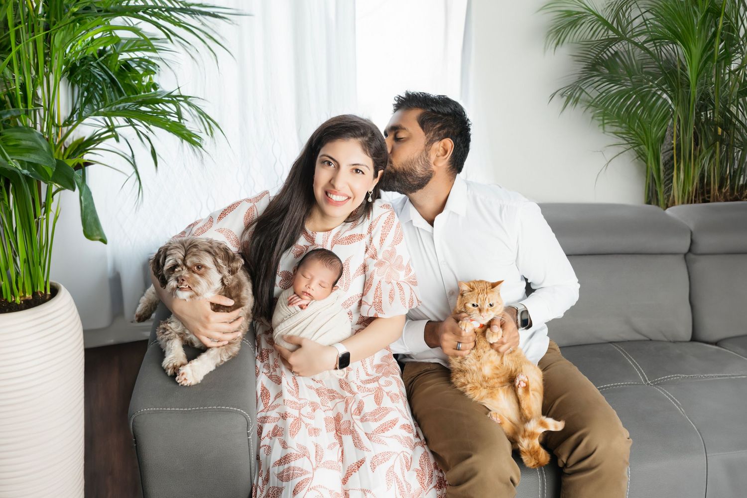 Family relaxing on grey couch with newborn baby and two cats in plant-filled living room.