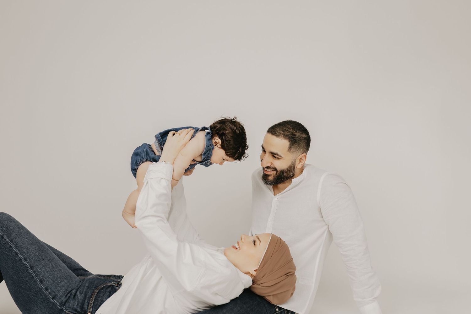 Muslim family portrait with father lifting baby, mother in hijab lying down, all wearing white in studio.