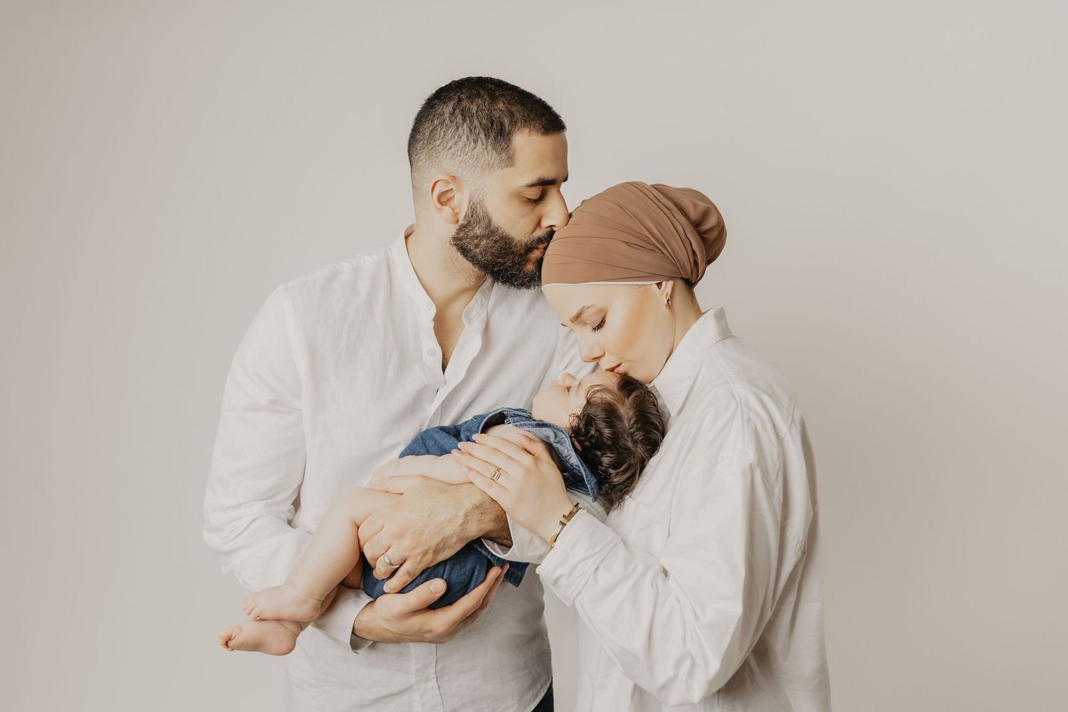 A couple in white shirts tenderly kisses while cradling their newborn baby, the mother wearing a tan head wrap.