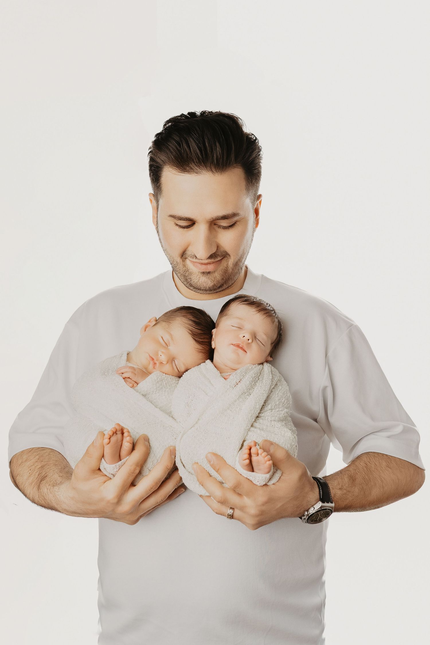 A smiling father in a white shirt holds two sleeping swaddled newborn twins against his chest.