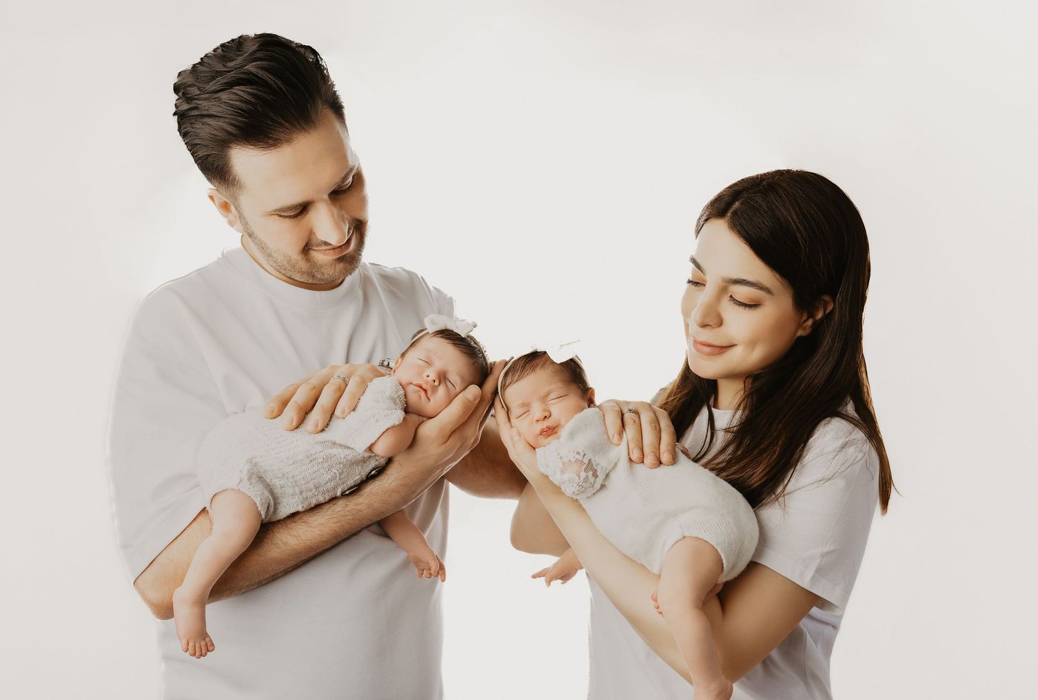 A young couple in white shirts tenderly hold their newborn twins against a white background.