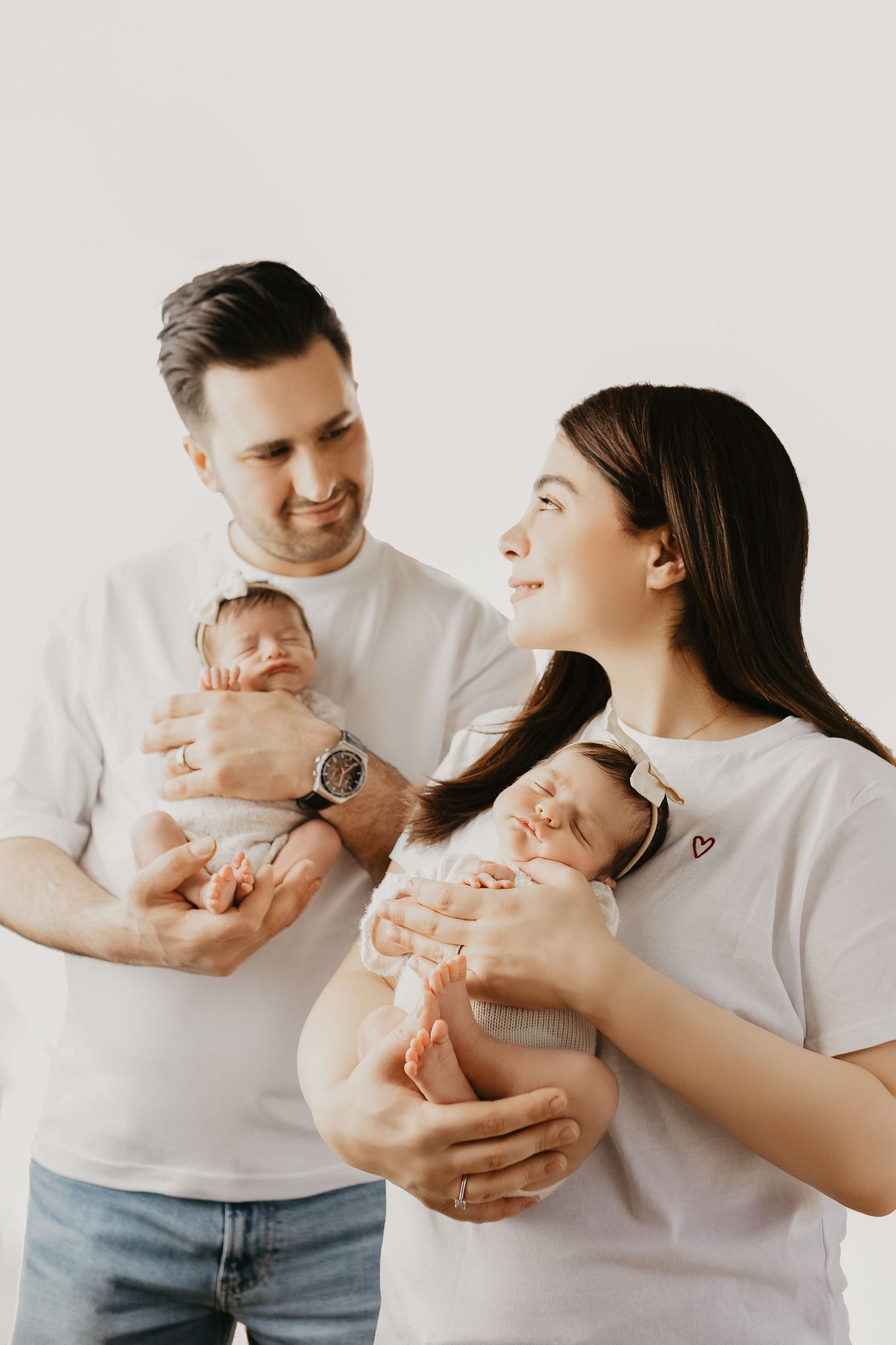 Happy parents in white t-shirts holding their newborn twins against a white background.