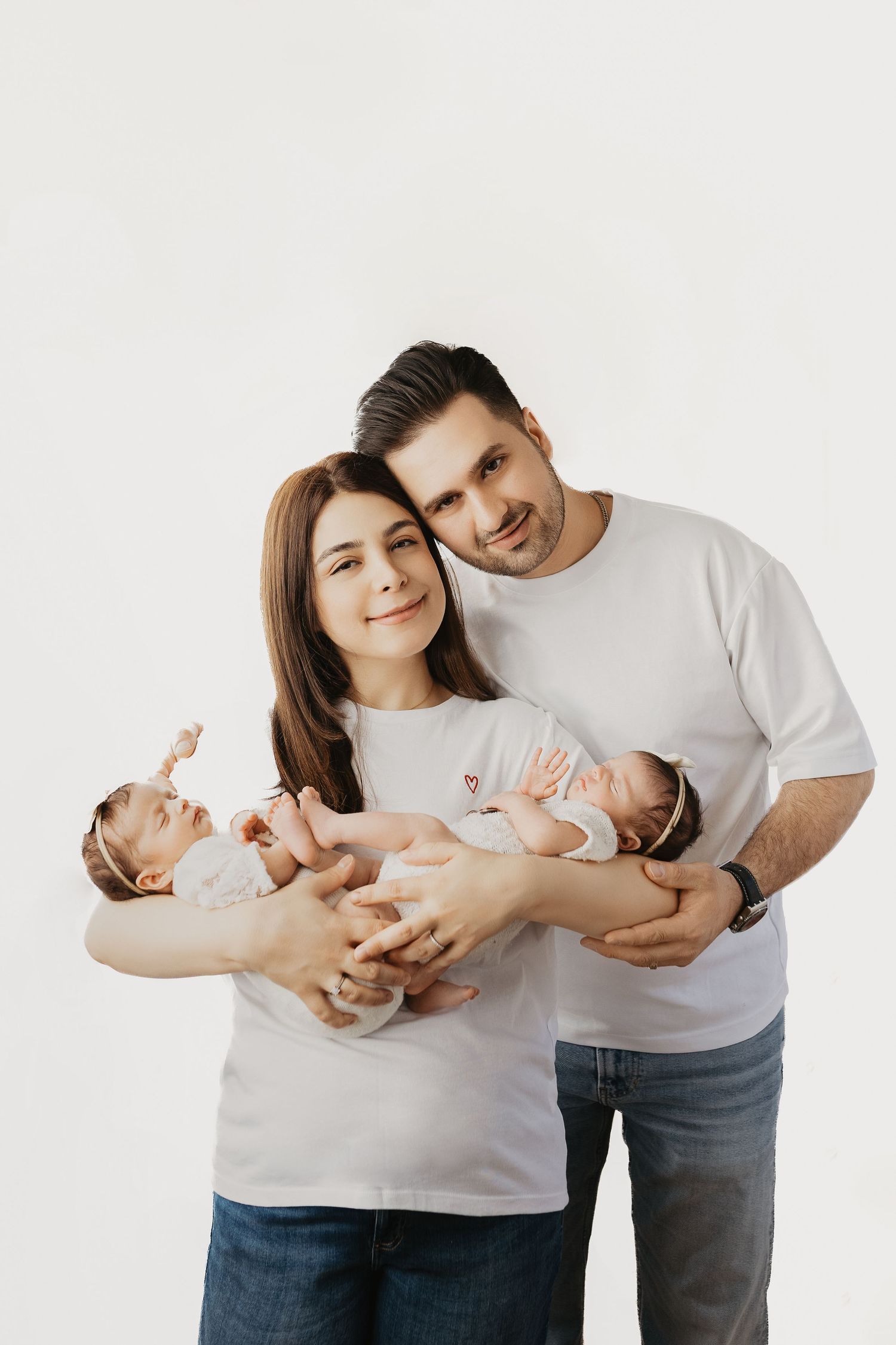 Happy parents in white t-shirts holding twin newborns, smiling at camera against white background.