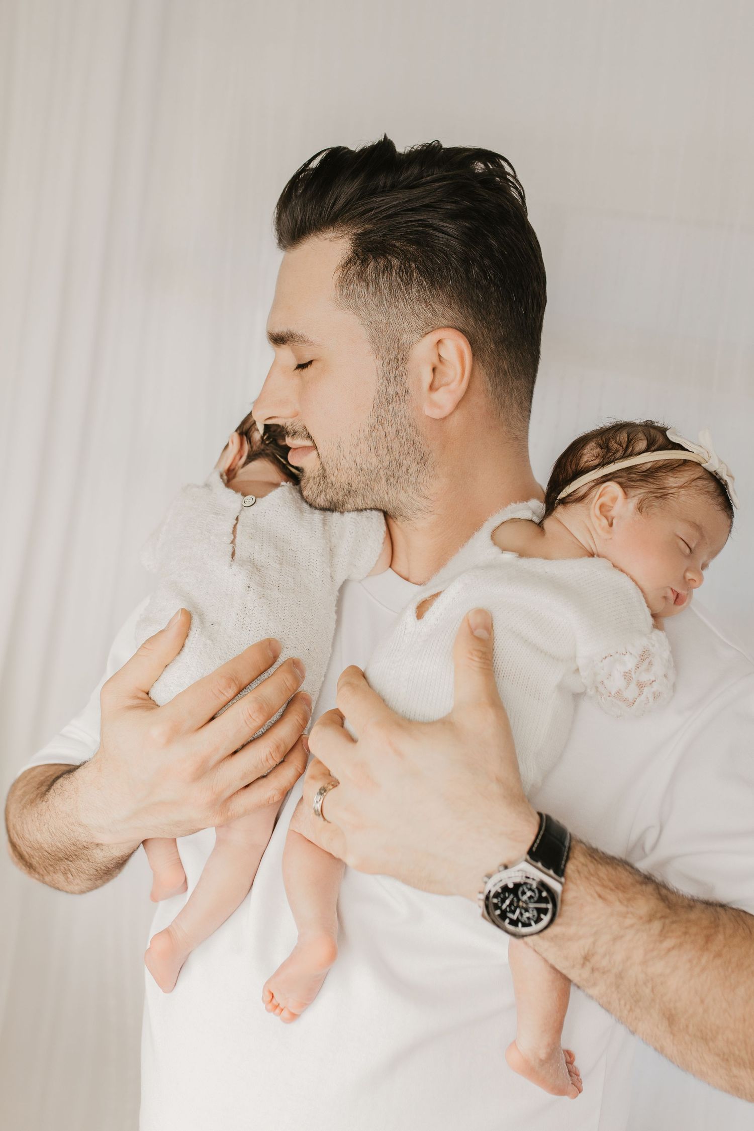 A smiling father in white holds his sleeping newborn baby girl dressed in white, wearing a watch.