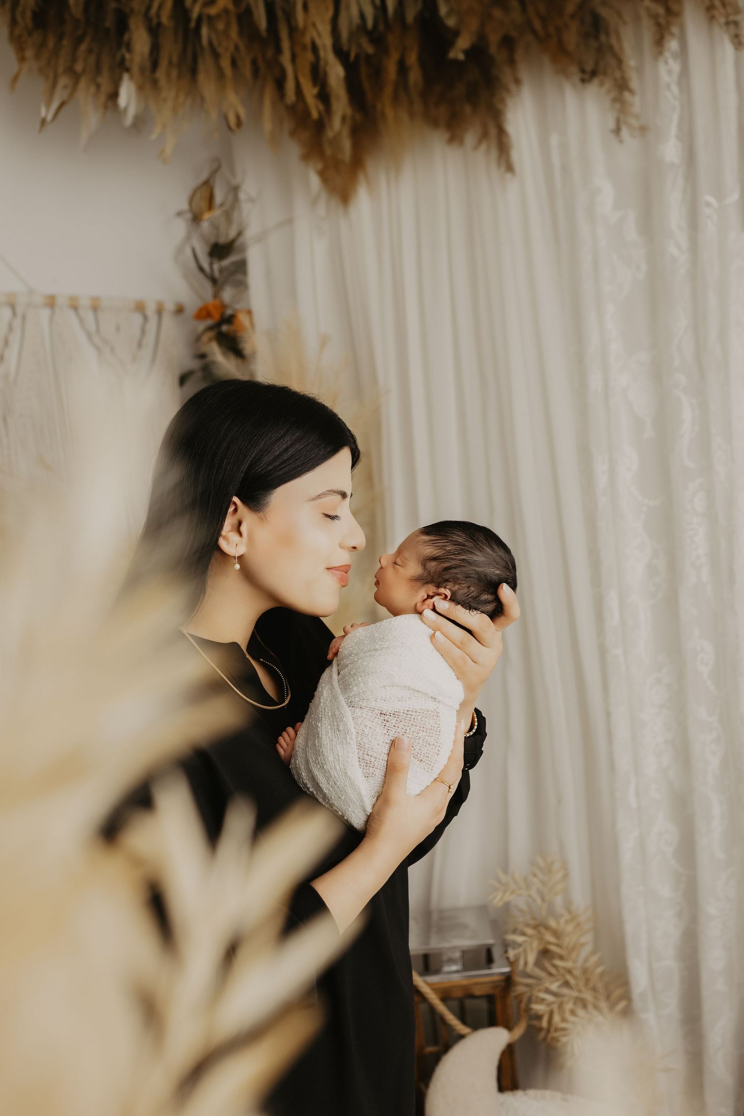 An intimate portrait of mother and newborn against white curtains with dried pampas grass decor.