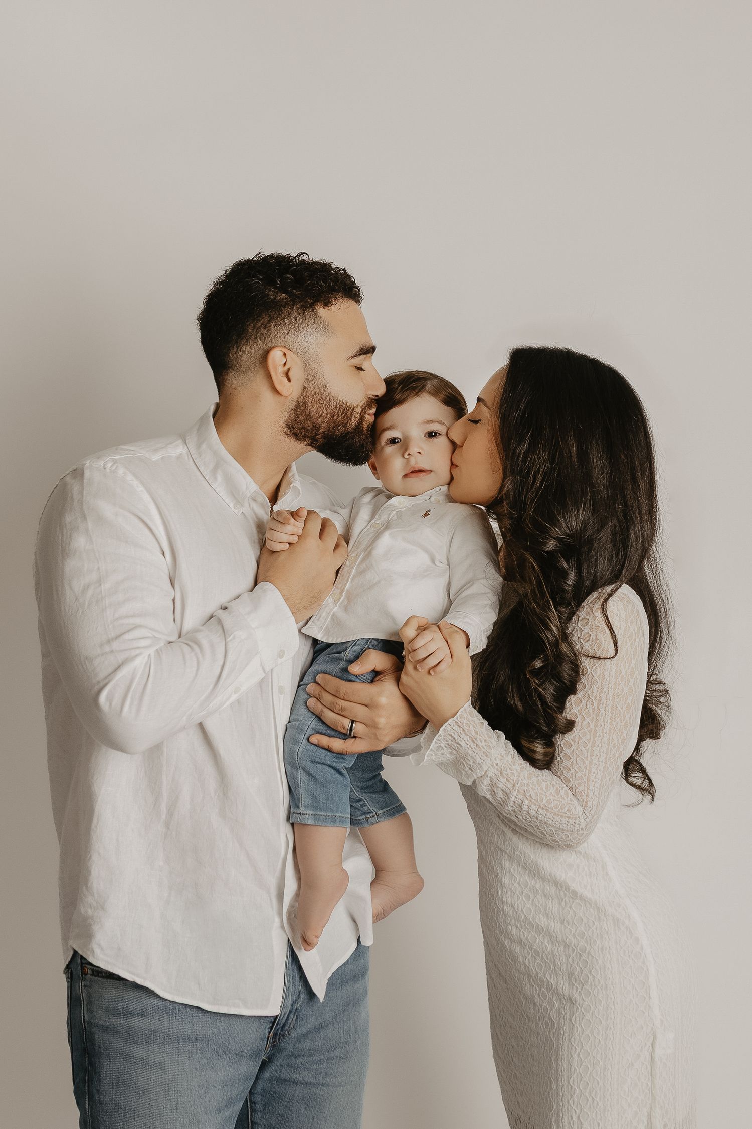 A couple in white outfits kisses their toddler on the cheeks while holding him against a neutral background.