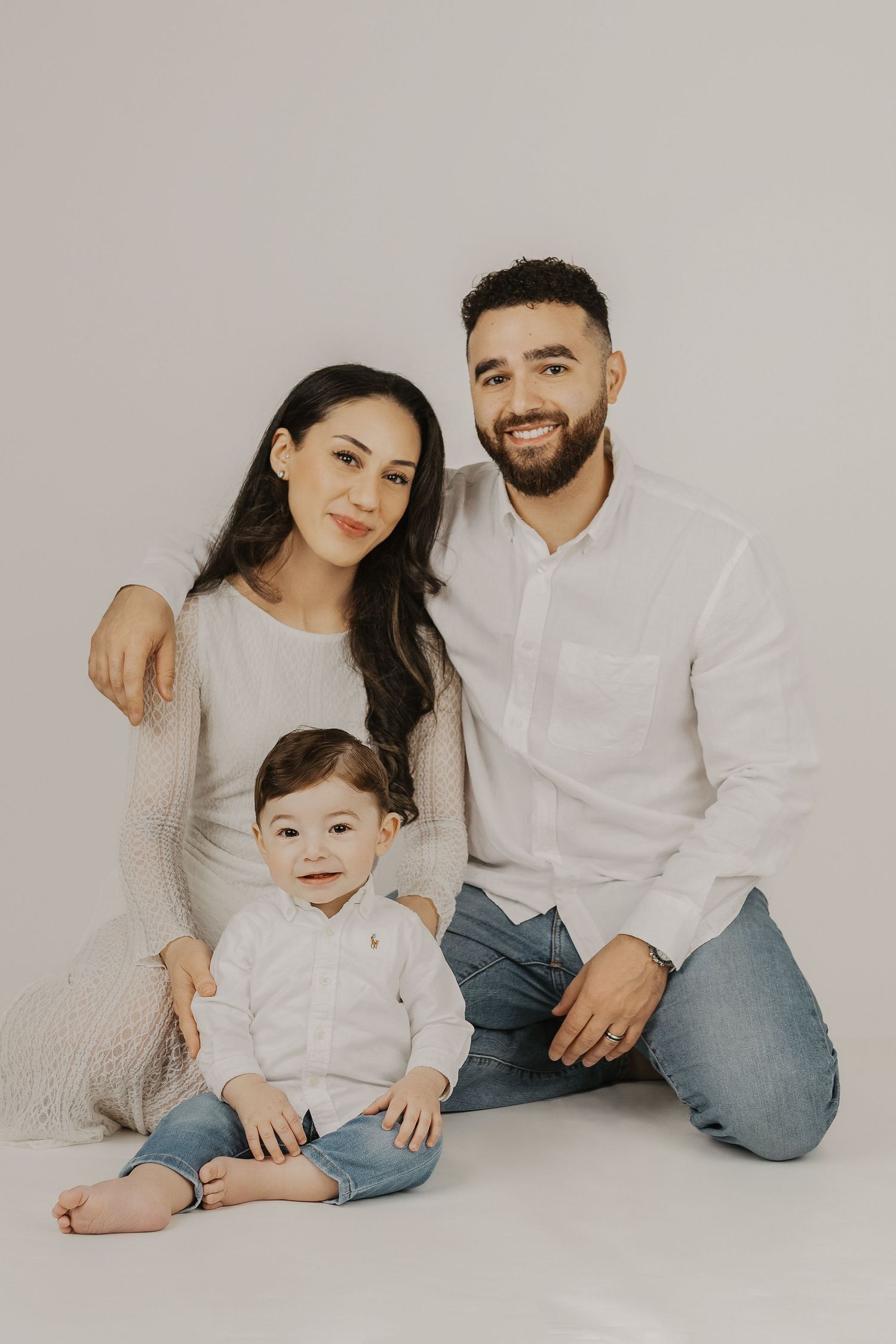 Young family portrait with smiling parents and baby in white outfits against a light background.