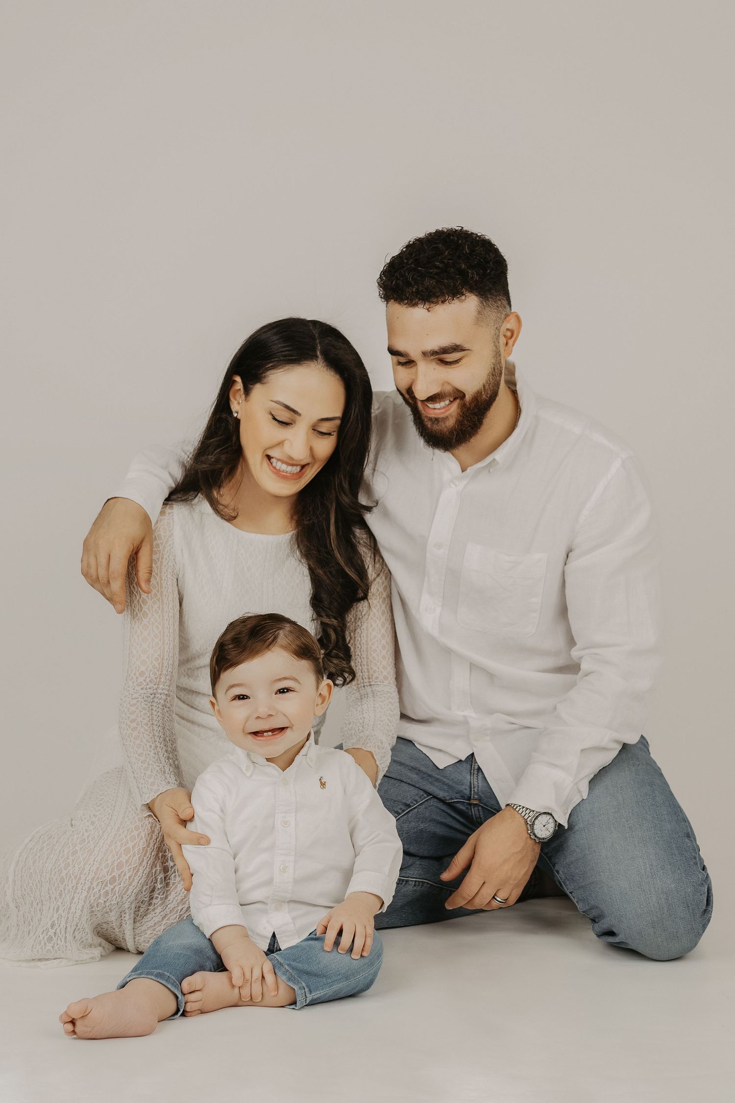 A happy young family with a baby boy poses together in white outfits for a studio portrait photo.