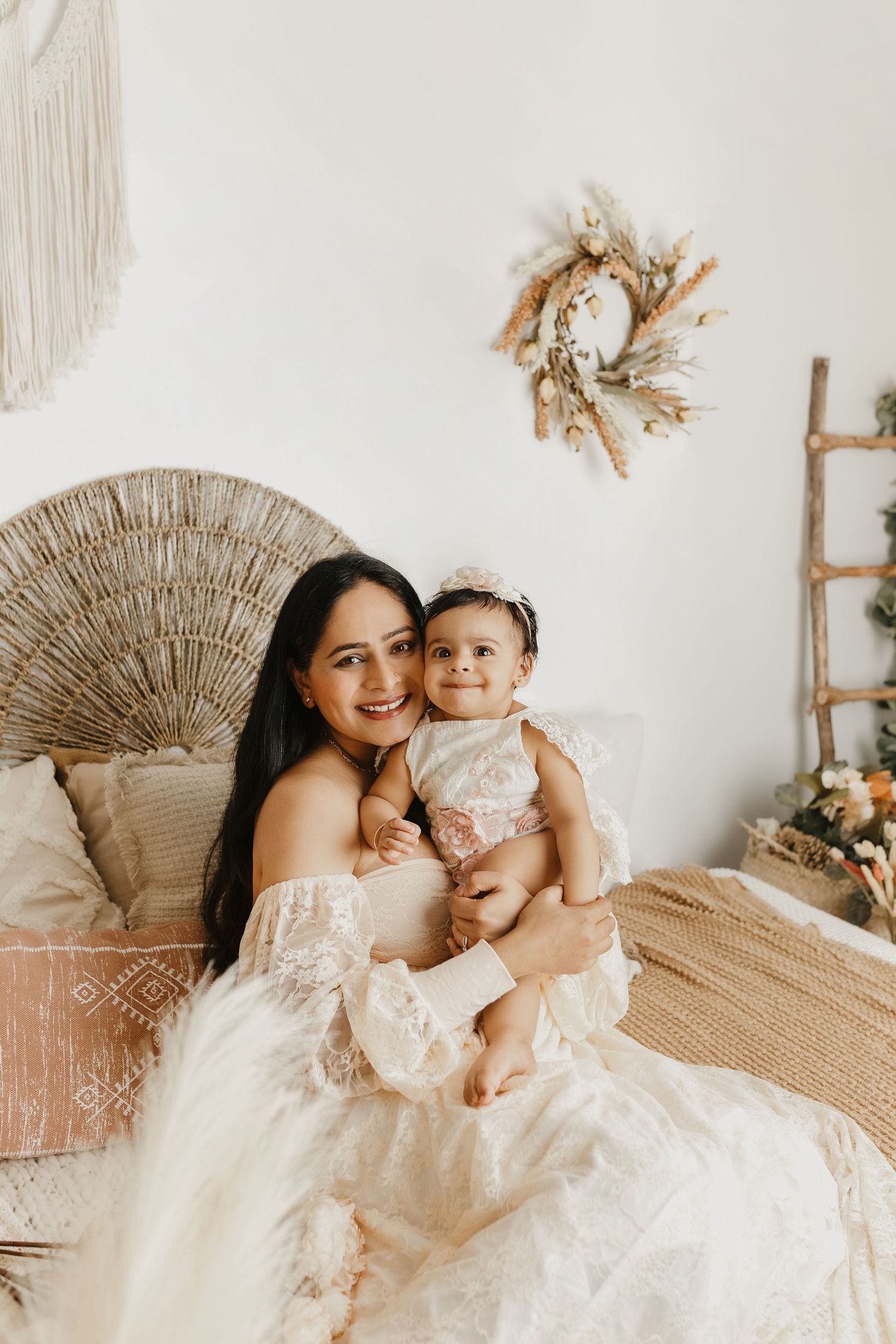 Mother and child wearing matching white lace dresses pose on a rattan chair with neutral decor.