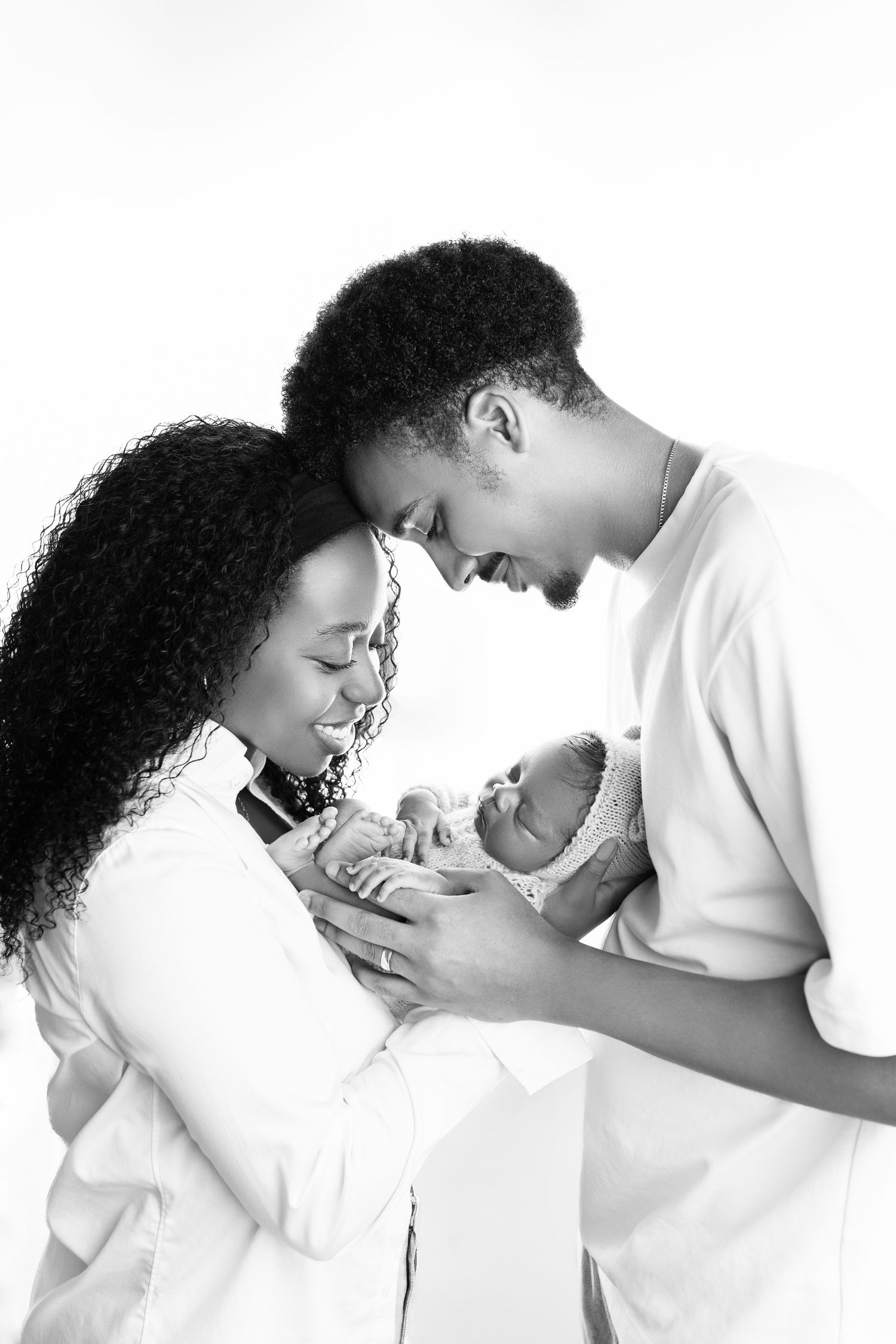 Black and white photo of smiling parents tenderly holding their newborn baby together against white background.