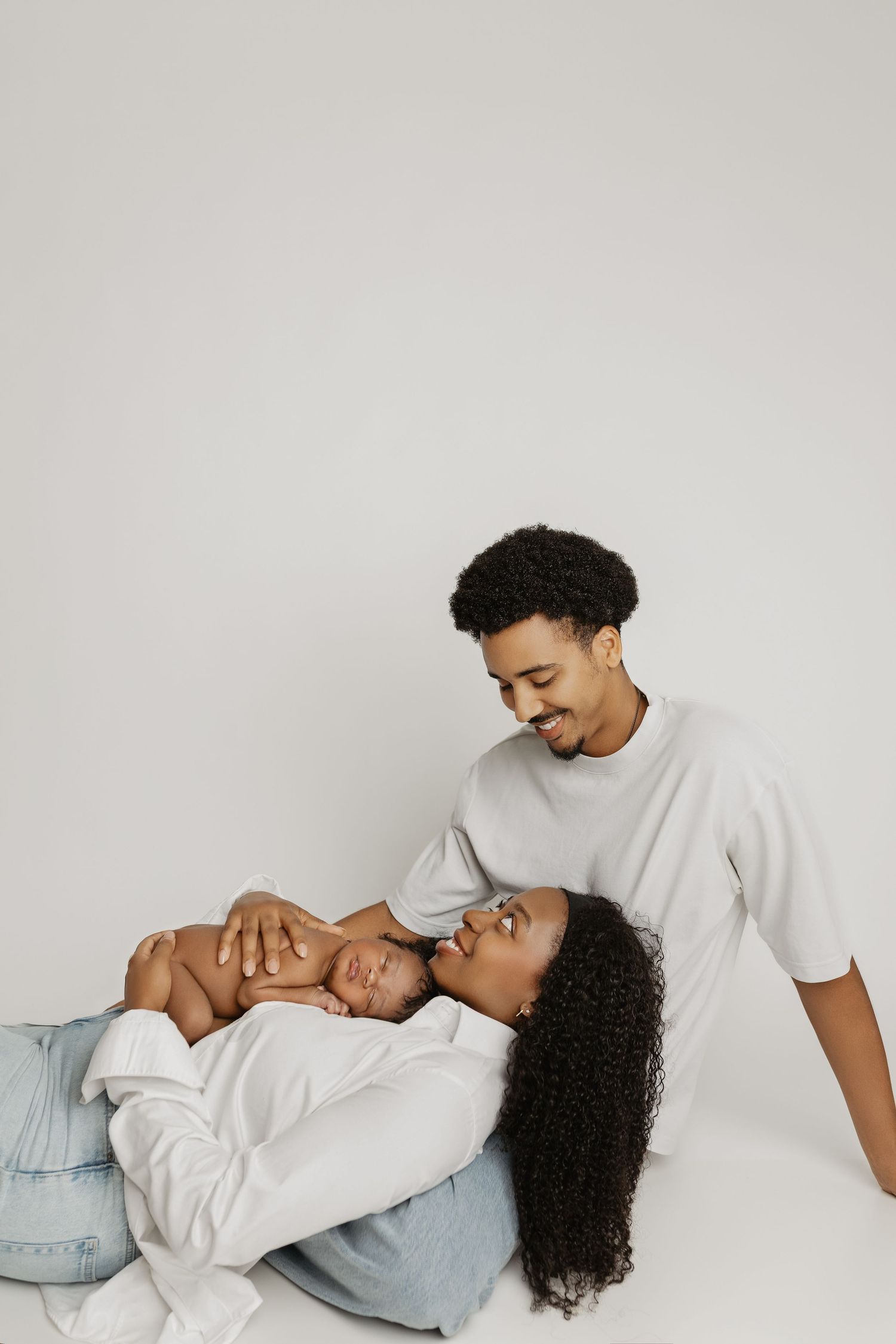 A happy Black family poses together, with mother lying down holding newborn baby while father smiles lovingly at them.
