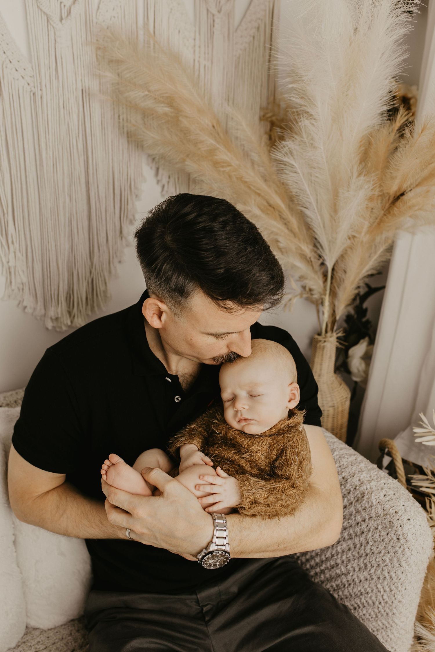 Family cuddle session on cozy bed with neutral decor and pampas grass in background.