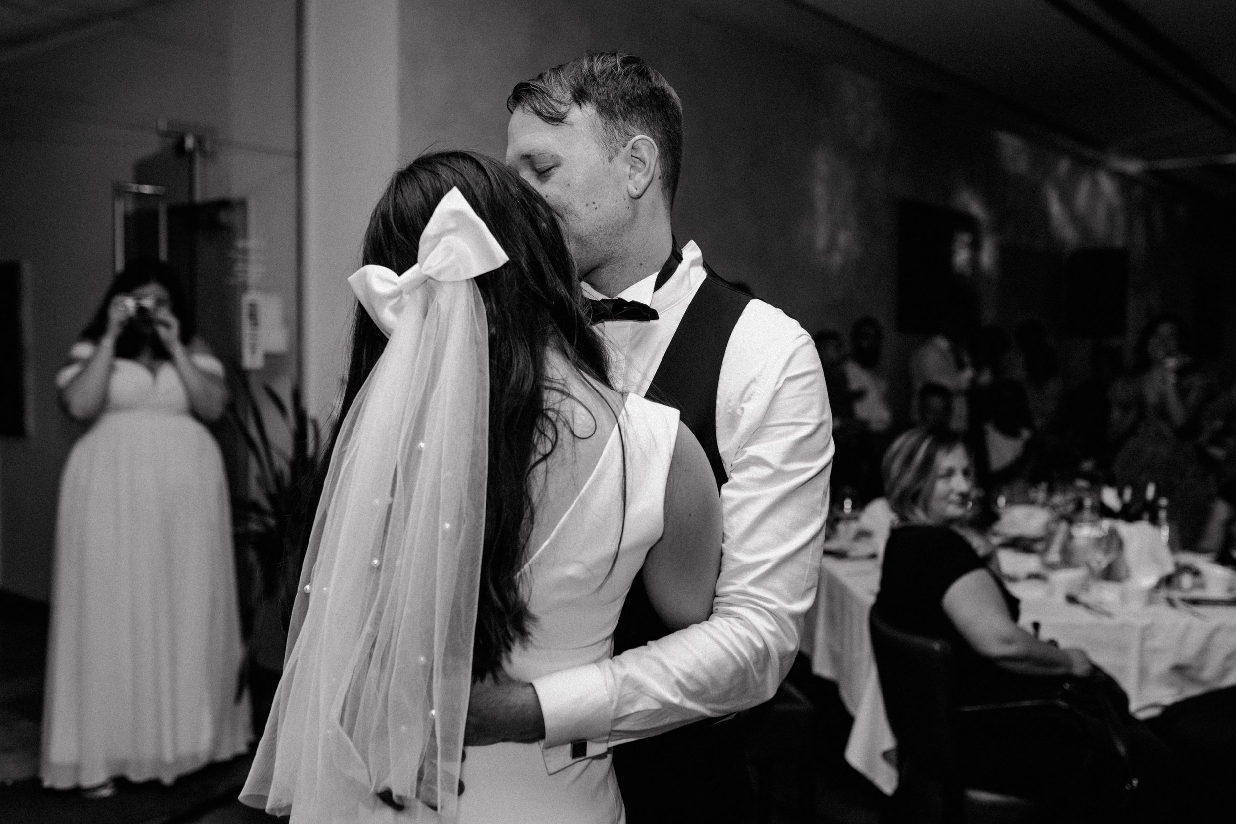 sweet moment of newlyweds kissing highlighting brides hair clip bow tie captured in black and white.