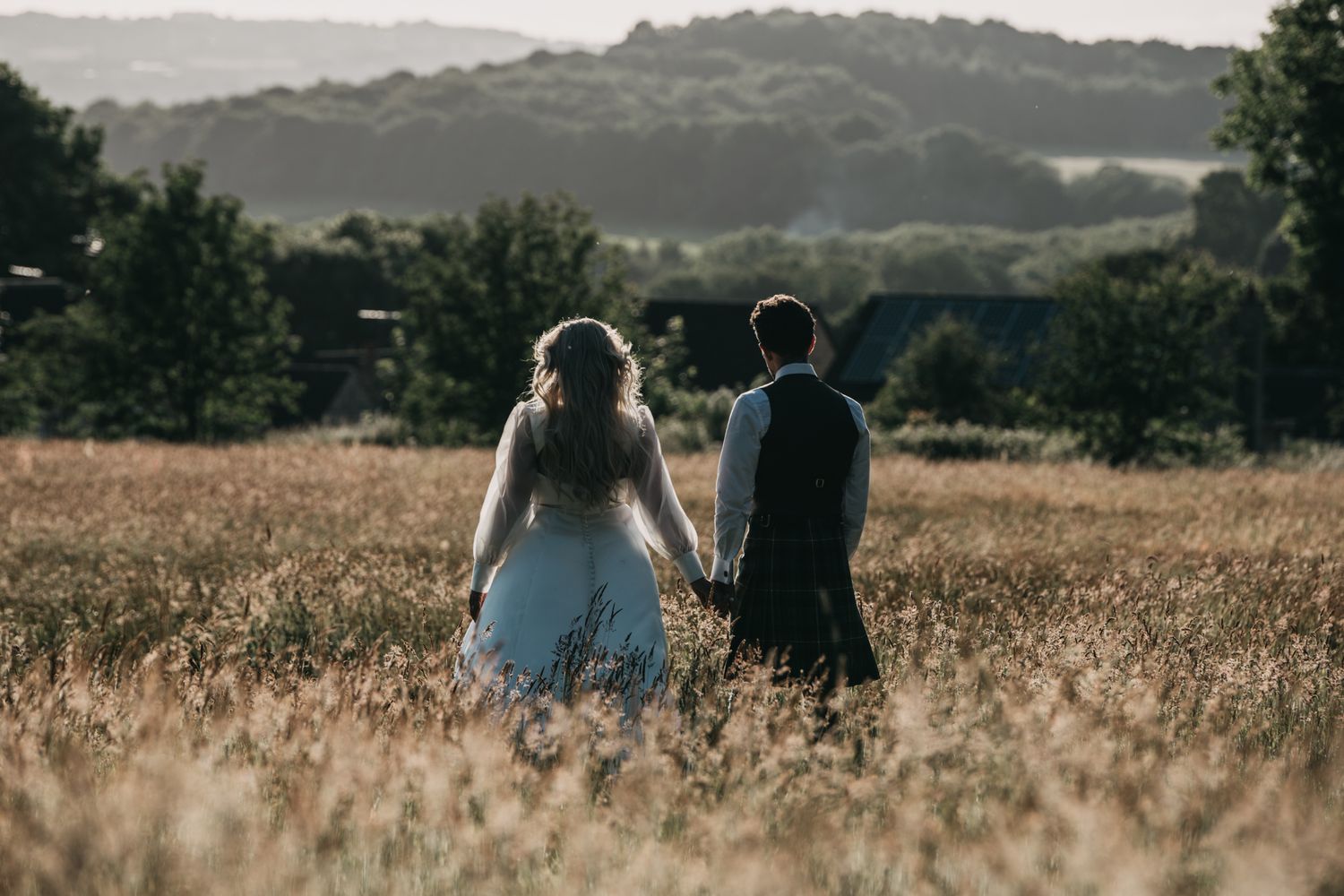 A newlywed couple stands in a golden field at sunset with mountains in the distance.