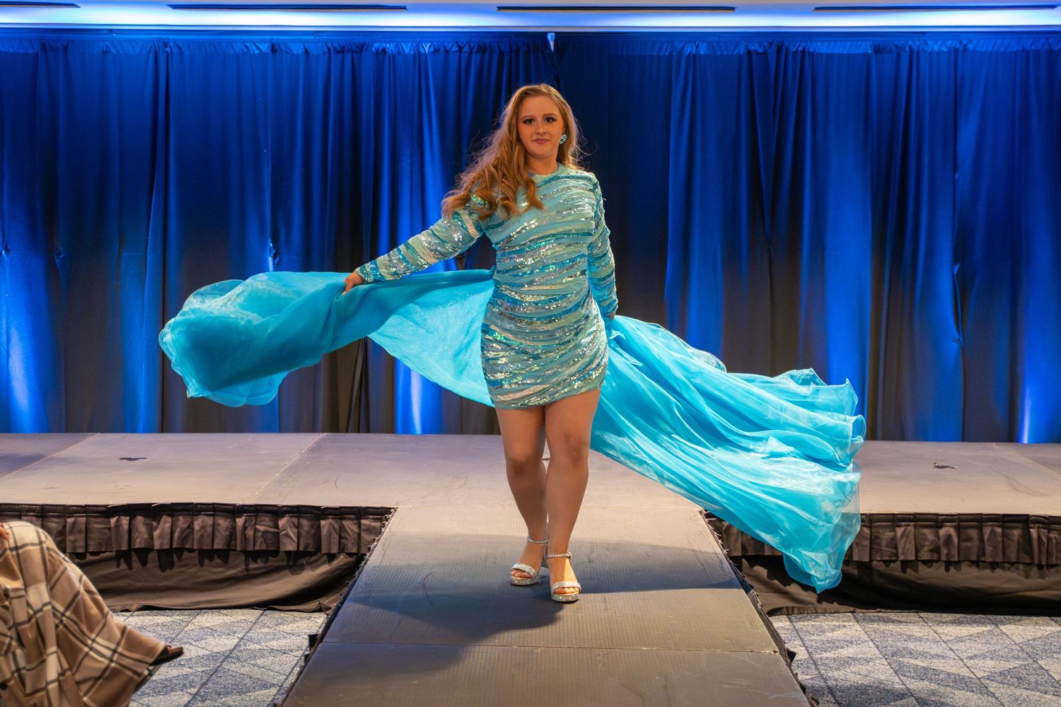 Fashion show on blue-lit runway featuring flowing turquoise evening gowns.