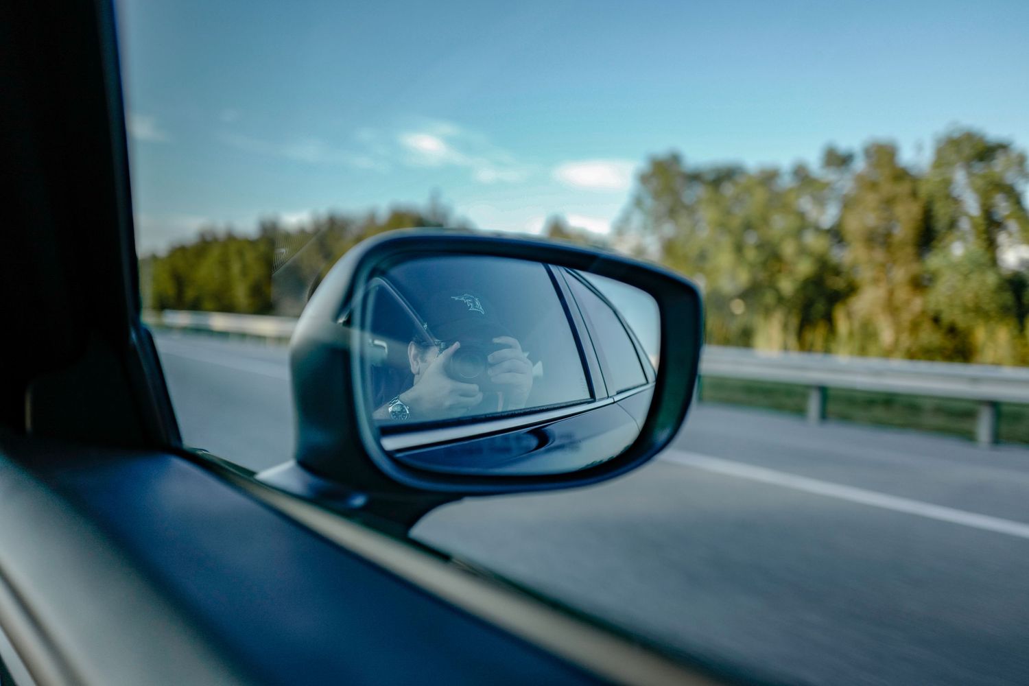 Side view mirror reflection on highway with blue sky and trees in background.