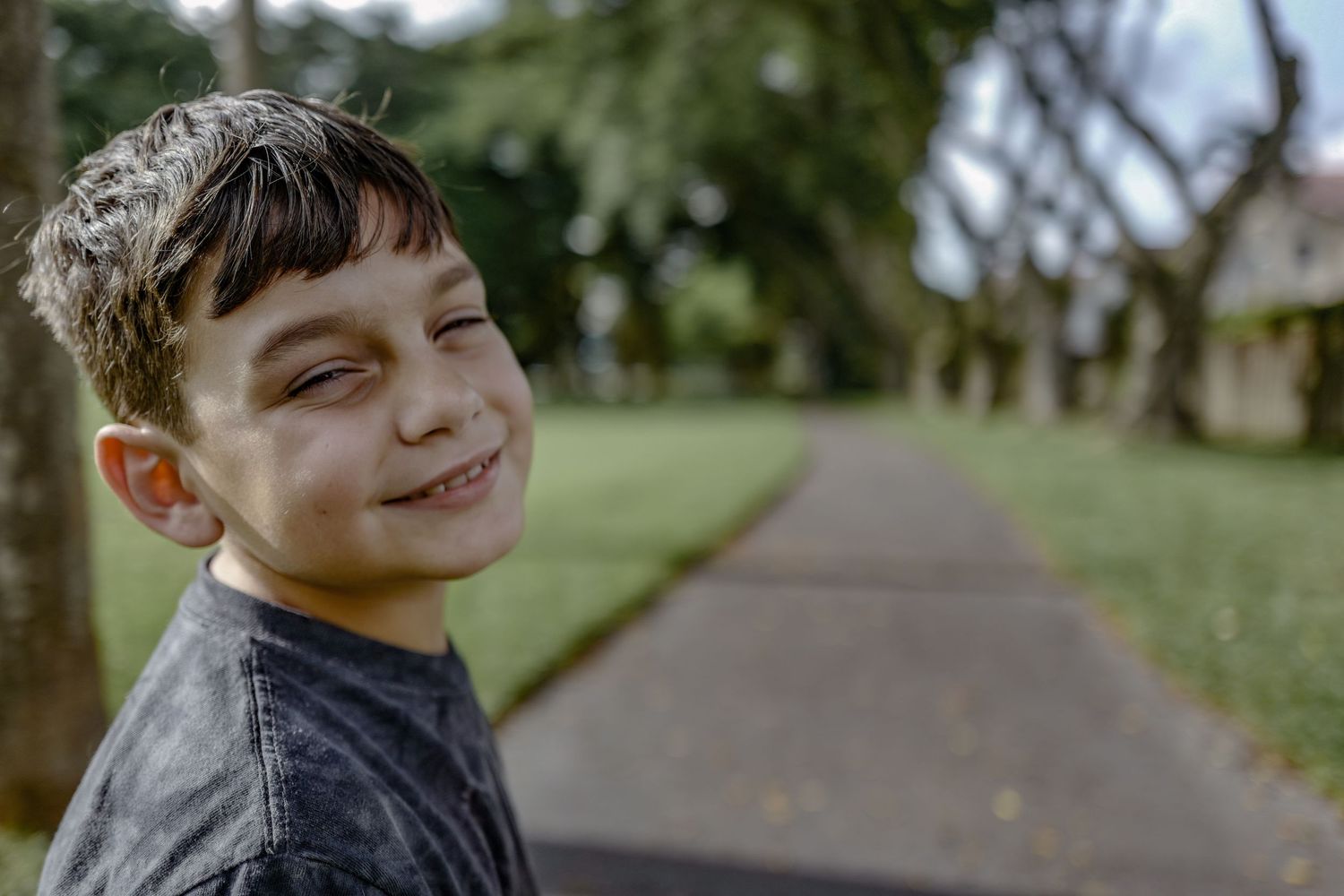 Young boy in gray shirt smiling warmly on tree-lined pathway.