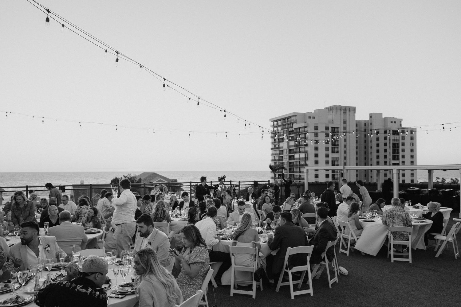 Outdoor rooftop wedding reception with string lights, guests dining with ocean view at dusk.