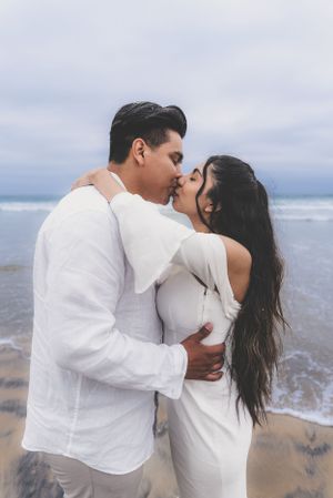 Couple sharing a passionate kiss on a sandy beach with ocean waves in the background during engagement session.