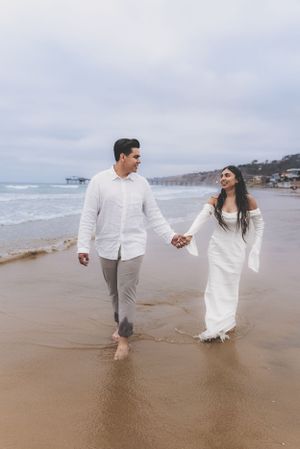 Smiling couple walking hand-in-hand along a wet sandy beach with a pier visible in the cloudy background.