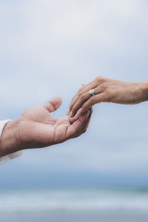 Close-up of two hands reaching toward each other, woman wearing a sparkling diamond engagement ring against a blue sky.