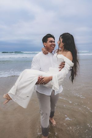 Groom carrying his laughing bride in his arms while standing in the ocean surf during a beach engagement session.