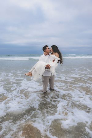 Couple kissing romantically as groom carries bride through ocean waves on a dramatic cloudy beach setting.