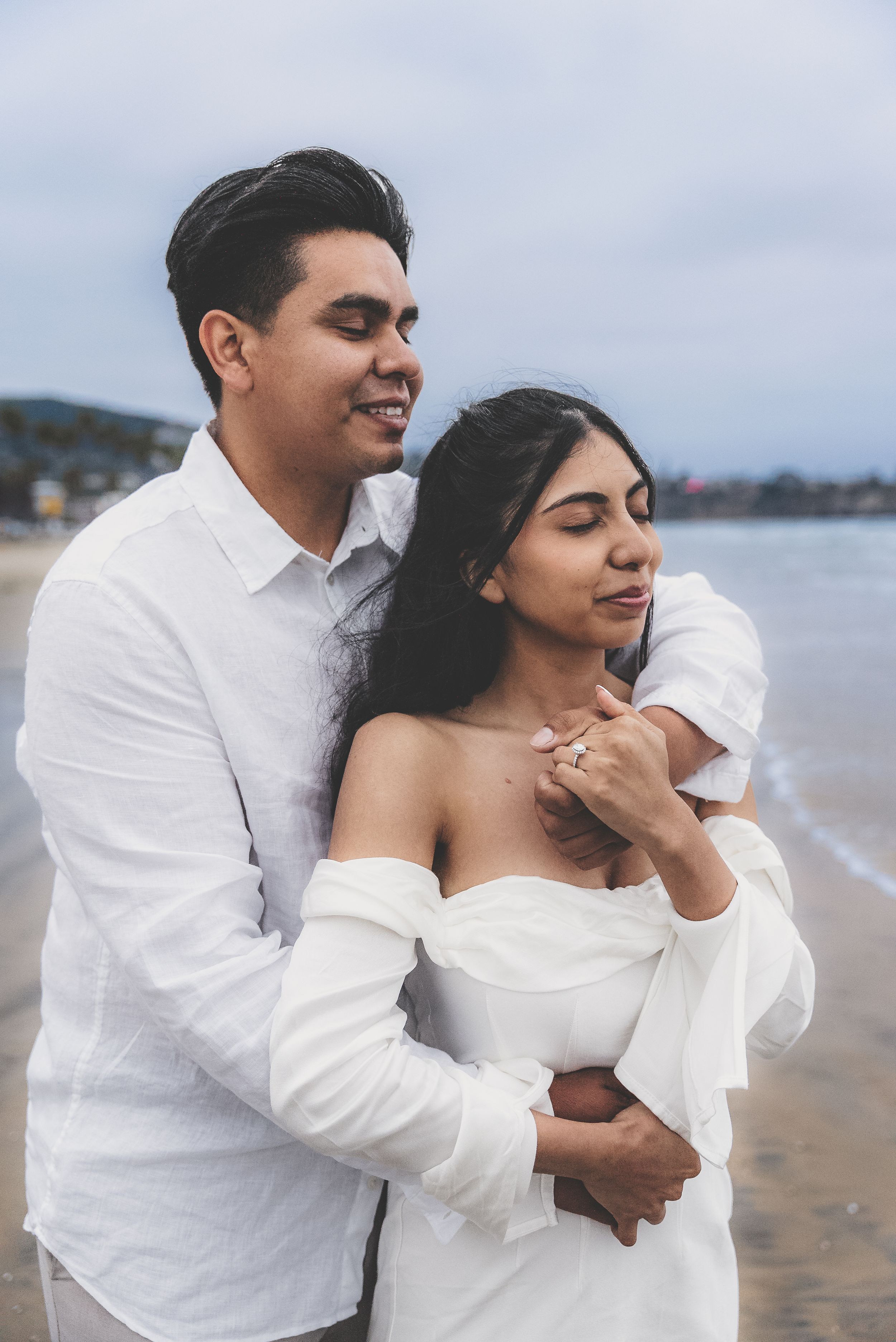 Couple embracing on beach in white outfits, man holding woman from behind near ocean shore.
