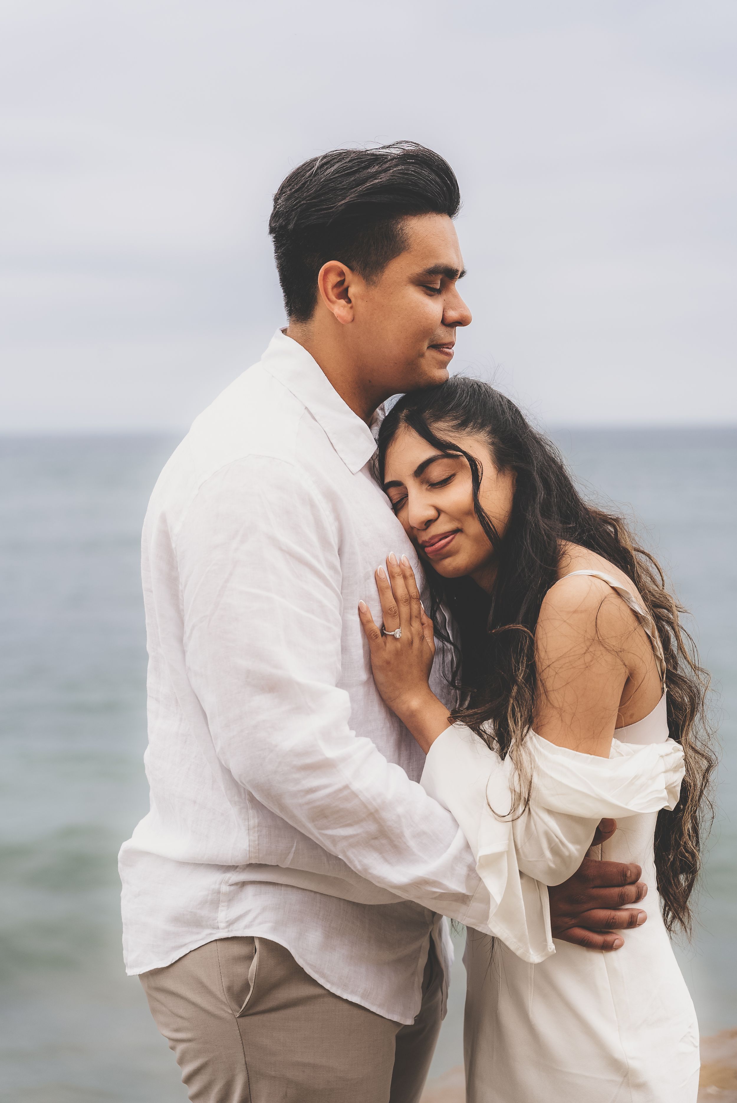 Romantic couple embracing on a cloudy beach, woman in white off-shoulder dress resting her head on man's chest.