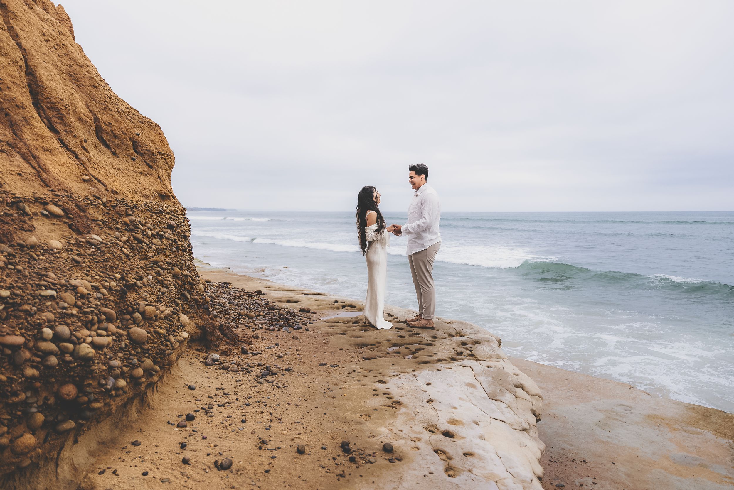 Engaged couple holding hands on rocky coastal cliffs with ocean waves crashing behind them during beach photo session.