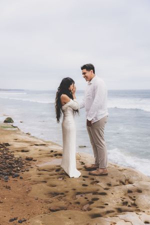 Happy couple sharing a joyful moment on a rocky beach, woman in elegant white dress laughing with her partner.