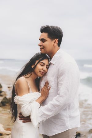 Couple sharing a tender embrace on a beach, woman in white off-shoulder dress leaning against man in white linen shirt.