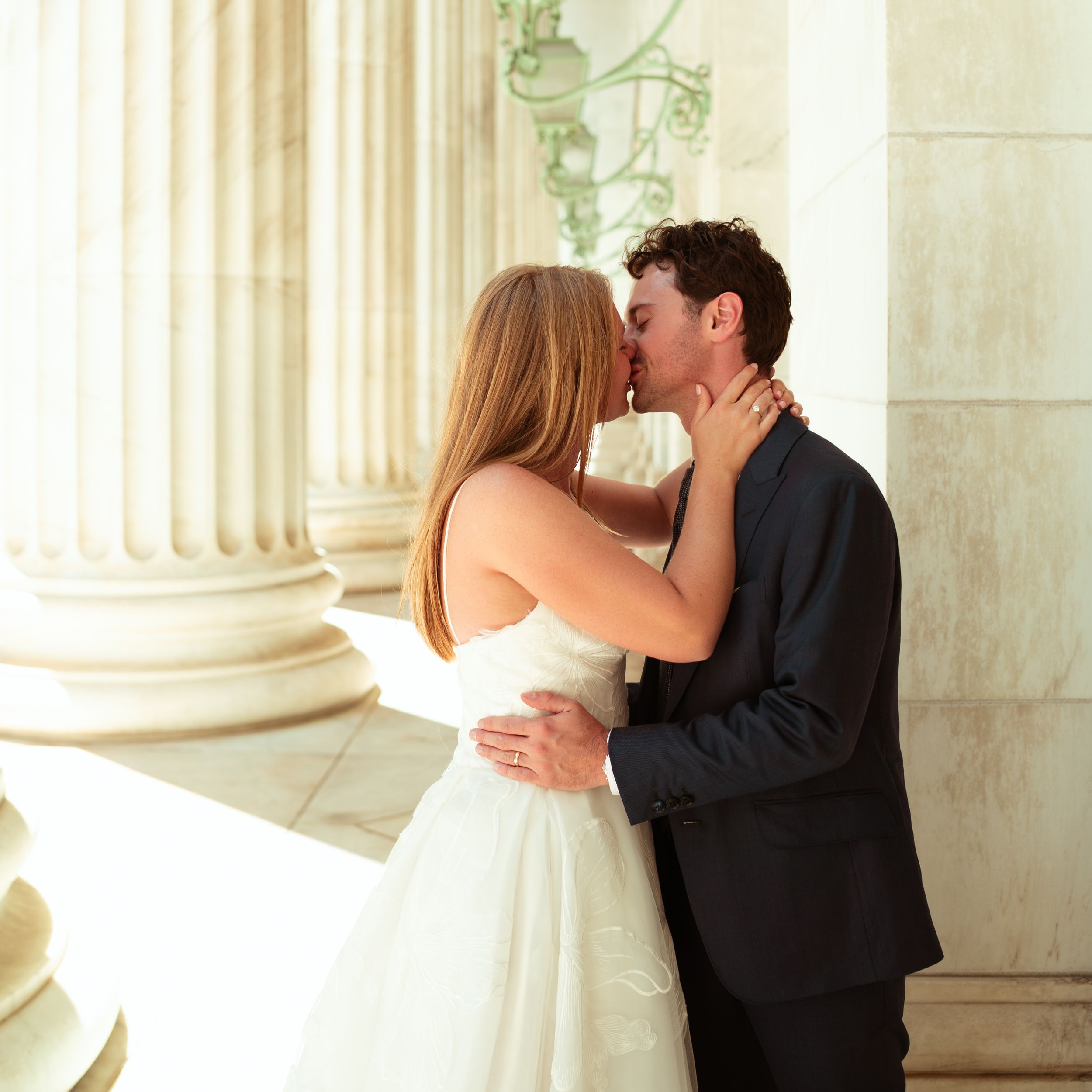 A romantic couple embraces and kisses in an elegant setting with white columns in the background.