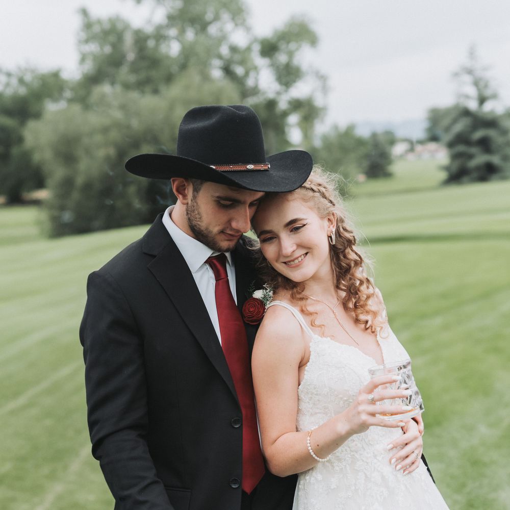Western themed wedding couple embraces outdoors on grassy hill wearing black cowboy hat and white dress.