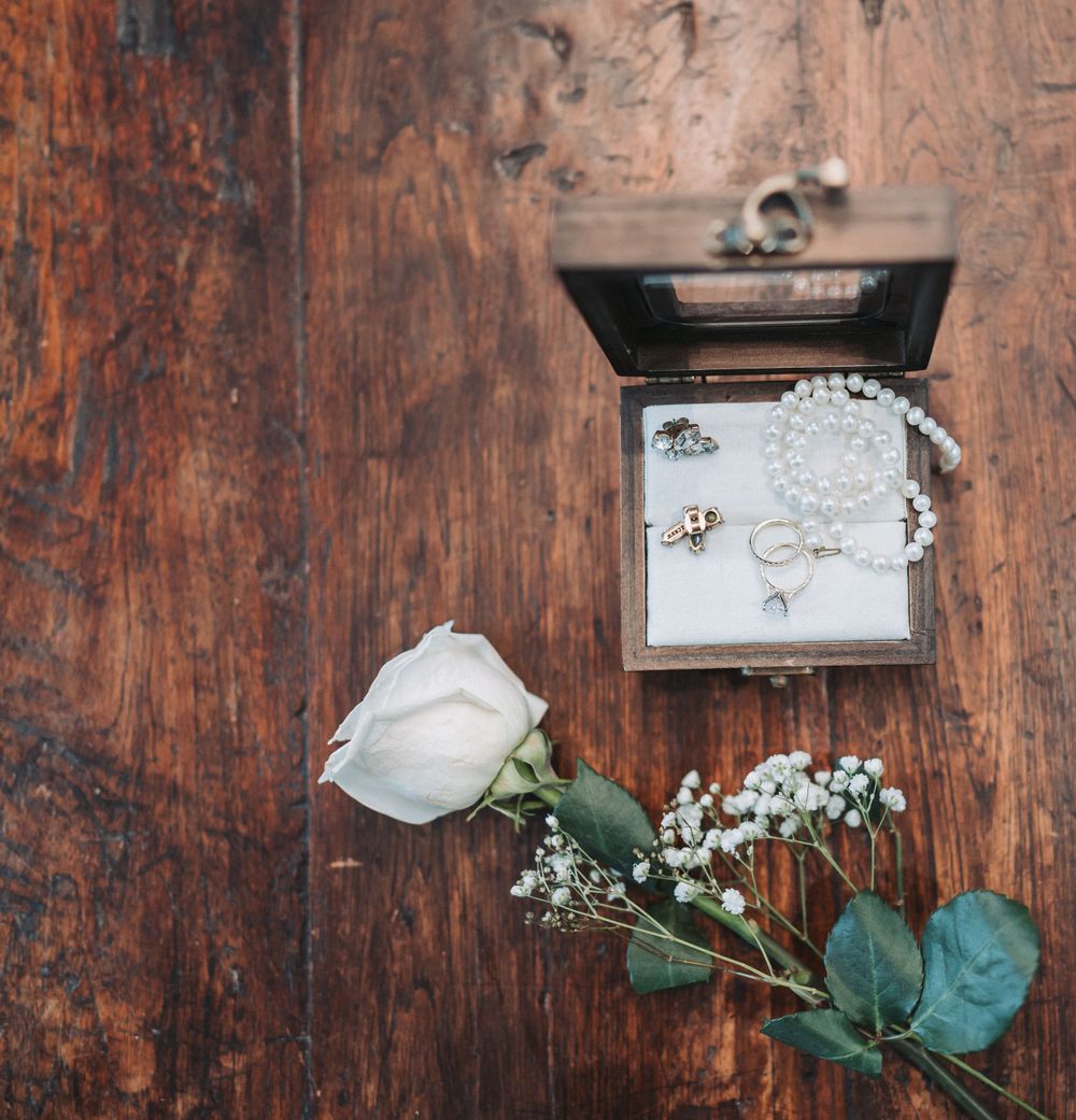 A delicate white rose and wedding rings displayed in an ornate wooden jewelry box.