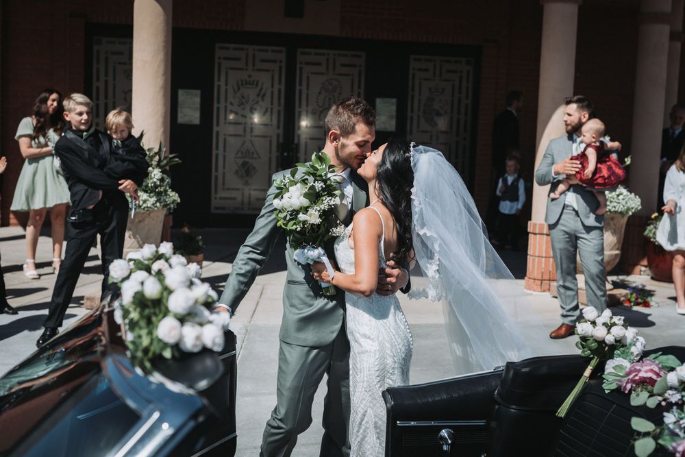 A romantic kiss between newlyweds during an outdoor wedding celebration with guests watching.