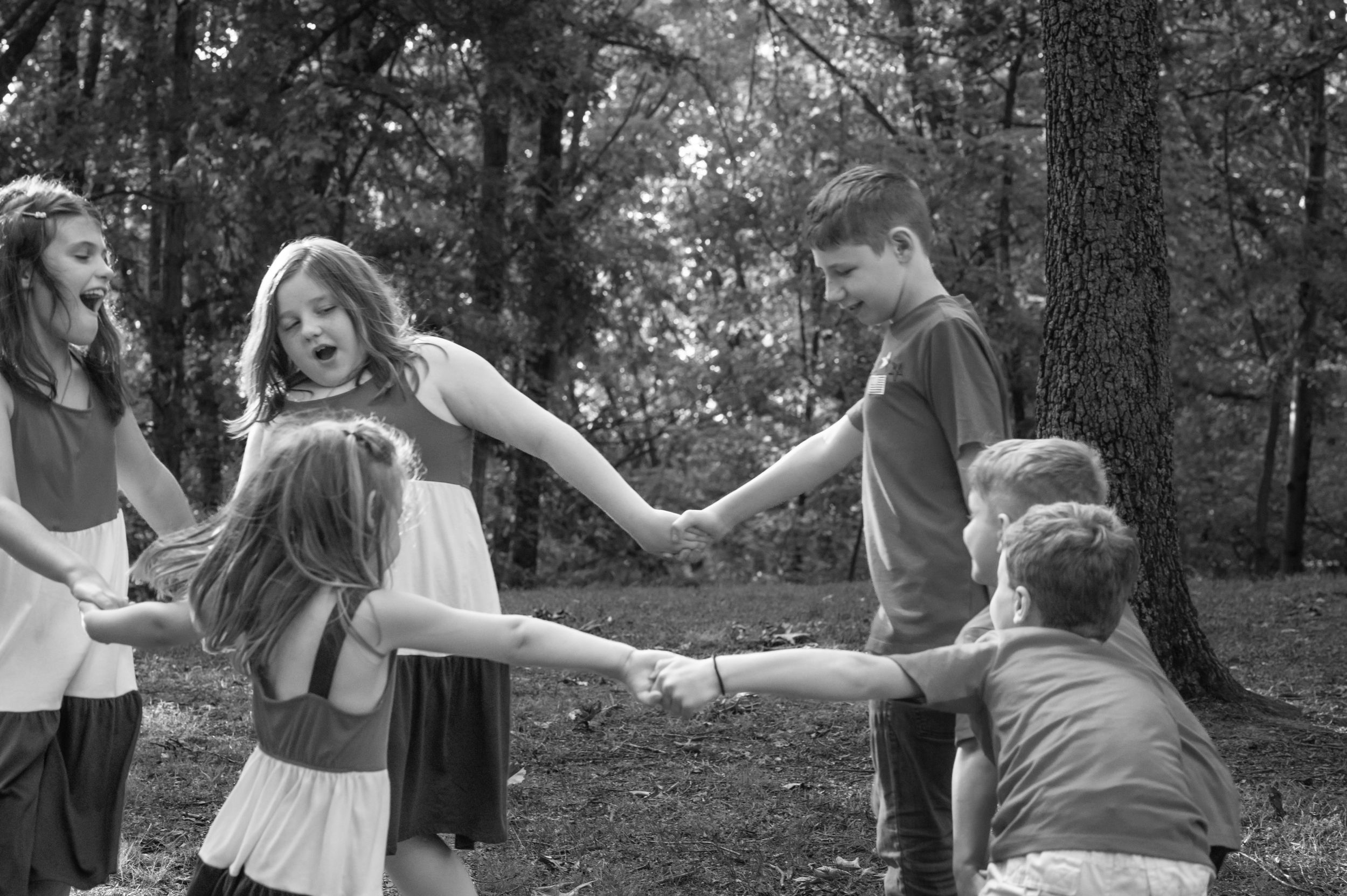 A group playing ring-around-the-rosie outdoors in a wooded setting.