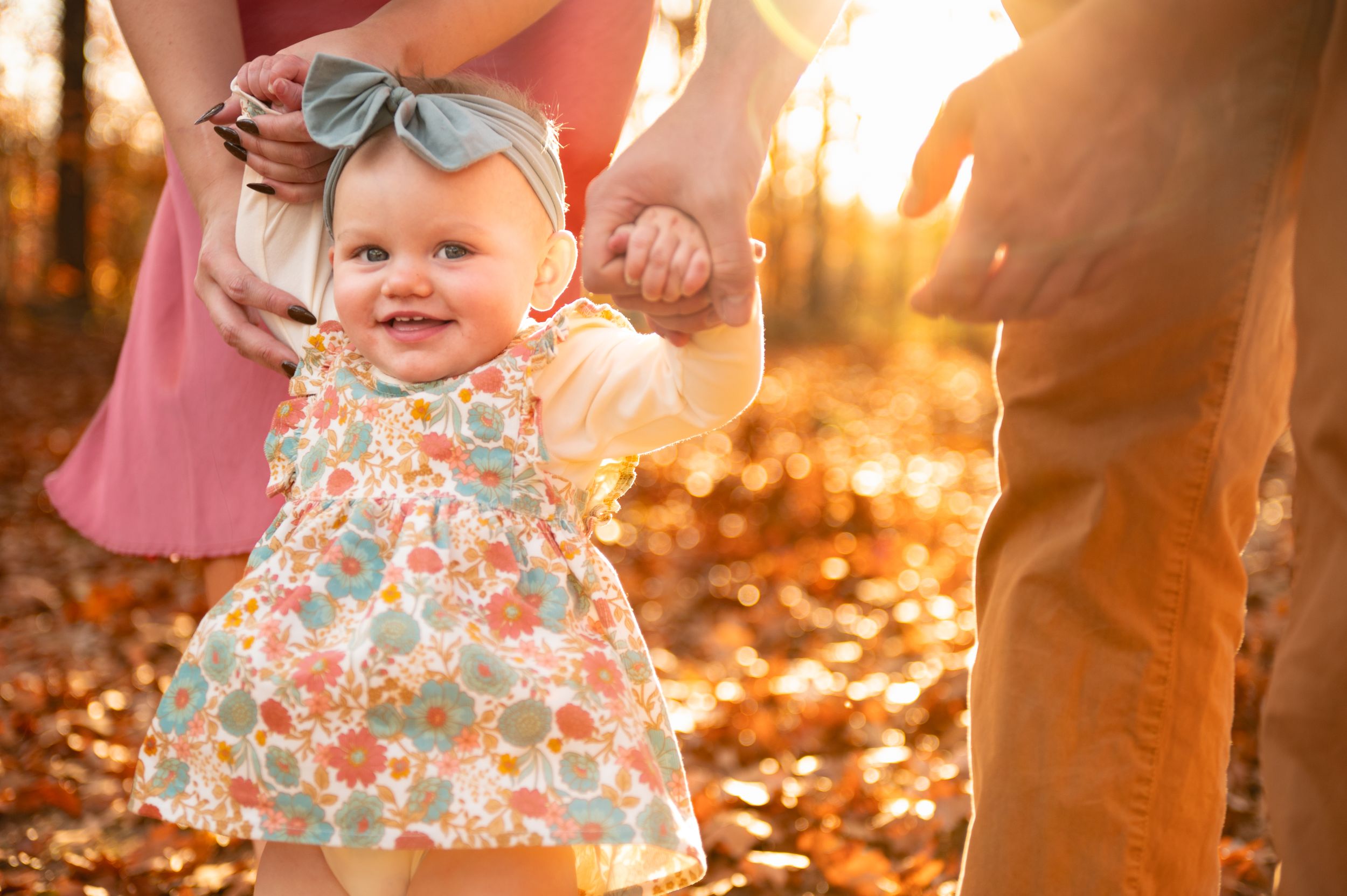 A toddler in a floral dress explores autumn leaves during golden hour.